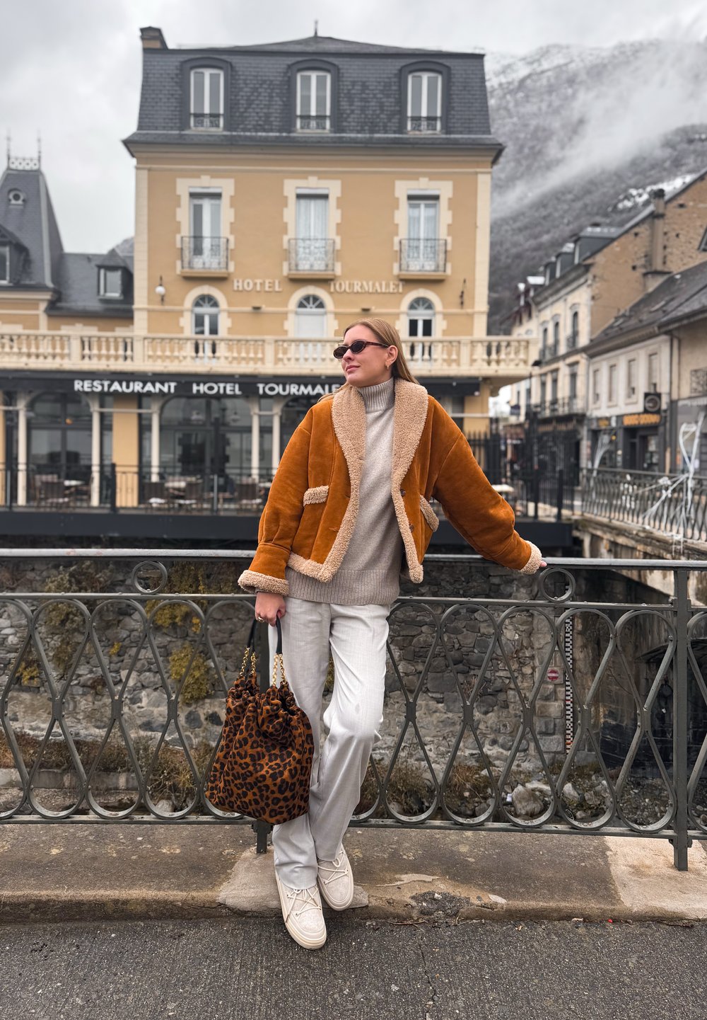 Γυναίκα με καφέ δερμάτινο μπουφάν με γούνα και λευκό παντελόνι στέκεται στη ράγα γέφυρας κρατώντας τσάντα με λεοπάρ σχέδιο, με το ξενοδοχείο Τουρμαλέ στο φόντο.