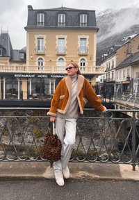 Γυναίκα με καφέ δερμάτινο μπουφάν με γούνα και λευκό παντελόνι στέκεται στη ράγα γέφυρας κρατώντας τσάντα με λεοπάρ σχέδιο, με το ξενοδοχείο Τουρμαλέ στο φόντο.