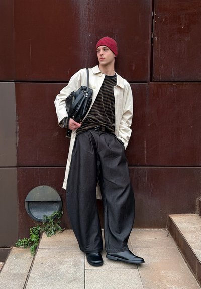 Persona joven apoyada en una pared marrón, vistiendo un gorro rojo, un abrigo beige, una camisa a rayas, pantalones anchos oscuros, botas negras y sosteniendo un bolso negro.