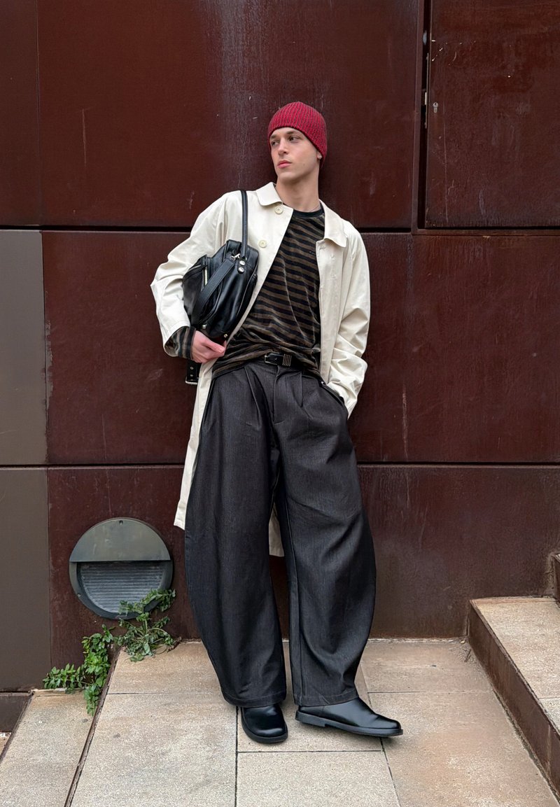 Jeune personne appuyée contre un mur marron portant un bonnet rouge, un manteau beige, une chemise rayée, un pantalon large foncé, des bottes noires et tenant un sac noir.