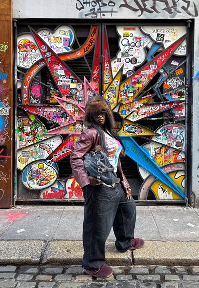 Mujer con gafas de sol, sombrero de piel, chaqueta de cuero y vaqueros anchos, de pie en una calle empedrada frente a una puerta metálica cubierta de grafitis coloridos.