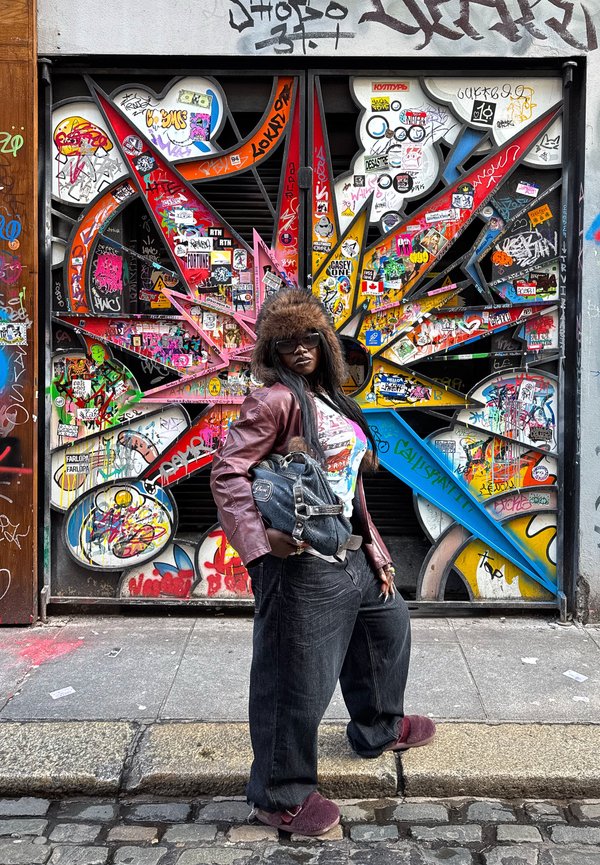 Kvinde med solbriller, pelshat, læderjakke og vide jeans står på brostensbelagt gade foran en farverig graffiti-beklædt metalport.