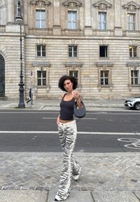 Canottiera nera attillata, pantaloni a zampa d'elefante grigio e bianchi a righe con un motivo testurizzato, con in mano una borsa a tracolla nera di forma rotonda. Pavimentazione in pietra e edificio sullo sfondo.