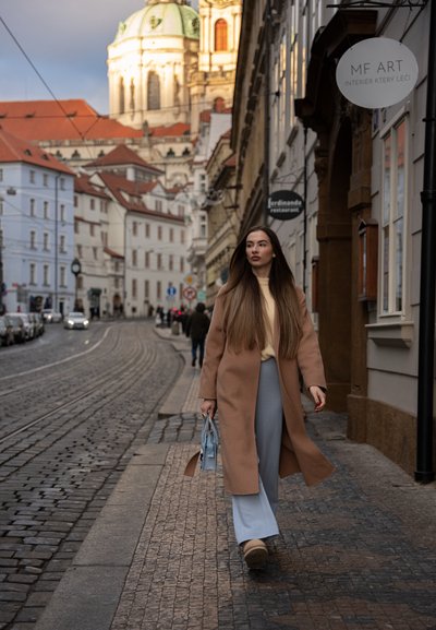 Mujer joven con abrigo beige y pantalones azules caminando por una calle empedrada con edificios históricos y una iglesia con cúpula al fondo.