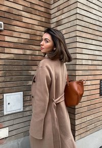 Femme aux cheveux mi-longs portant un manteau beige et un sac à main en cuir marron, debout près d'un coin de mur en briques et regardant en arrière.