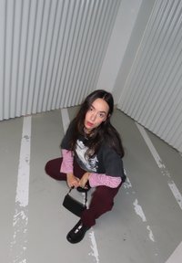 Femme accroupie dans un coin d'une zone de stationnement, vêtue de hauts superposés noirs et roses, pantalon bordeaux, bottes noires, tenant un petit sac à main noir.