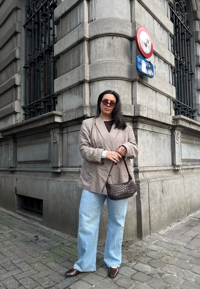 Mujer con blazer beige de gran tamaño y vaqueros azul claro sosteniendo un bolso marrón oscuro de pie en una esquina de una acera de la ciudad.