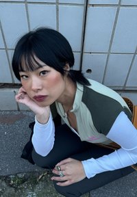 Jeune femme aux cheveux noirs attachés, portant des hauts blancs et verts superposés, assise à l'extérieur sur le trottoir près d'un mur carrelé, regardant vers la caméra.