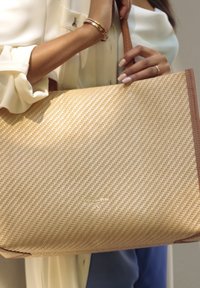Beige geflochtener Einkaufstasche mit braunen Lederakzenten, die eine strukturierte Oberfläche und ein markantes Logo aufweist. Die Tasche wird von einer Hand gehalten.