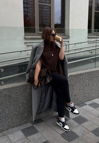 Cappotto grigio su un maglione a coste marrone, leggings neri e sneakers bianco e nere. Tiene una tazza di caffè, con una borsa marrone accanto a lei.