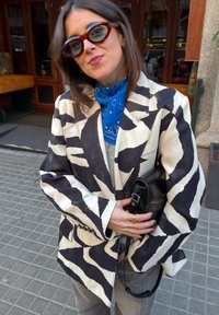Femme portant un blazer oversize à rayures zébrées, un foulard bleu noué autour du cou, des lunettes de soleil, tenant un sac à main noir sur un trottoir en ville.