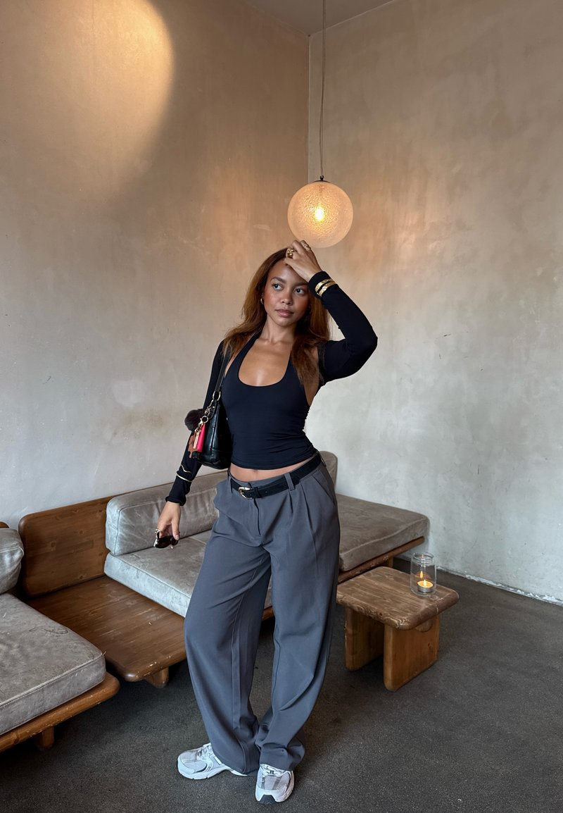 Frau mit braunen Haaren, die ein schwarzes Oberteil und graue Hosen trägt, steht in einem minimalistischen Wohnzimmer mit Holzmöbeln und einer hängenden runden Lampe.
