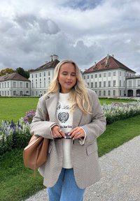 Beige ruudullinen ylisuuri bleiseri valkoisen tekstipaidan päällä, yhdistetty vaaleansinisiin farkkuihin. Ruskea käsilaukku ja hopeiset sormukset; taustalla kukkia ja rakennus.