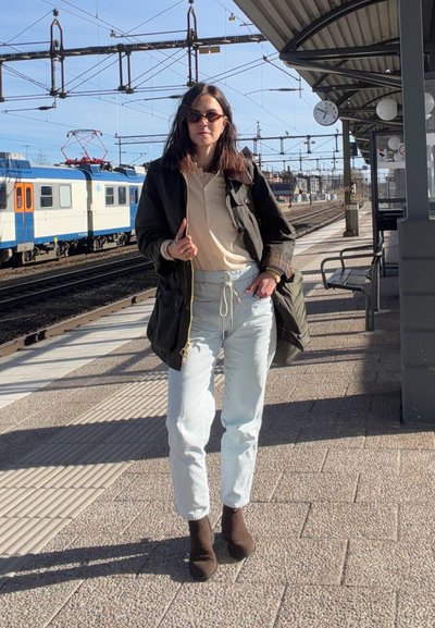 Mujer con gafas de sol, chaqueta negra, camiseta beige y pantalones blancos de pie en un andén de tren soleado con un tren azul y blanco al fondo.
