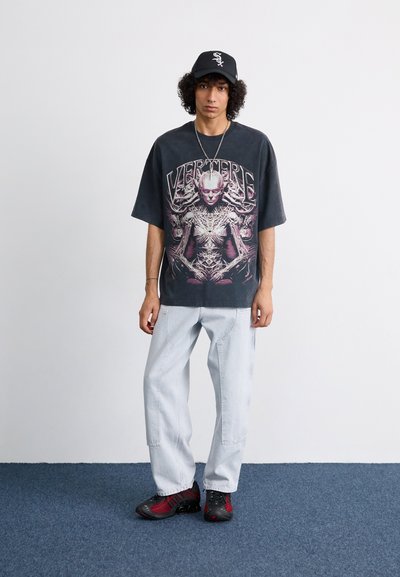 Camiseta negra oversized con un detalle en rosa, pantalones vaqueros baggy de color azul claro y zapatillas deportivas negras y rojas, todo sobre un fondo neutro.