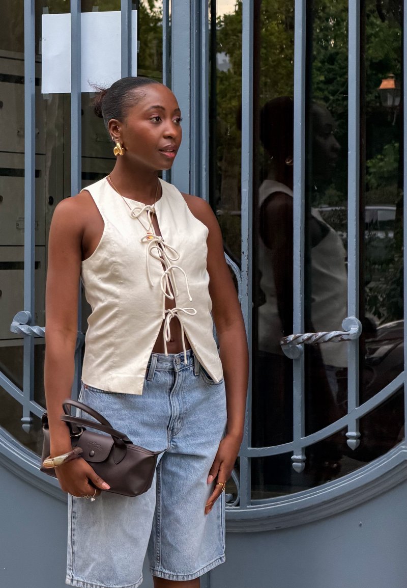 Frau mit dunkler Haut, trägt ein ärmelloses cremefarbenes Top und helle Denim-Shorts, hält eine kleine braune Handtasche und steht bei einem grauen Metalltor.