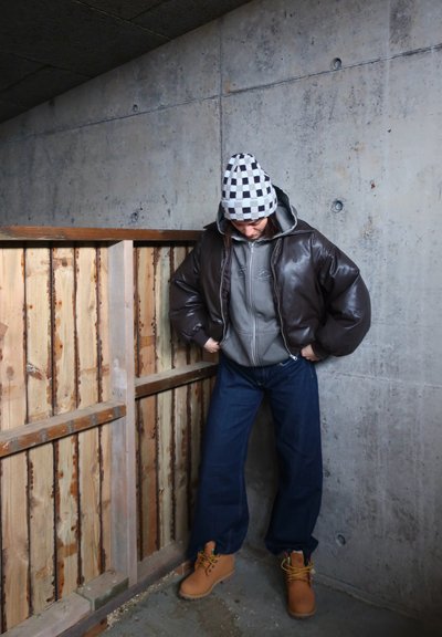 Chaqueta bomber acolchada marrón, sudadera con cremallera gris, vaqueros anchos azules y botas de trabajo color marrón claro. Gorro de cuadros en blanco y negro. Fondo de valla de madera.