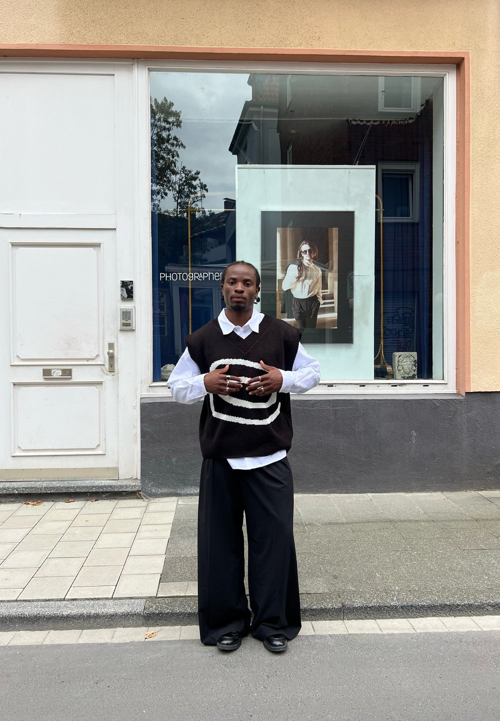 Una persona è in piedi davanti a un negozio, indossando un gilet a righe bianche e nere sopra una camicia bianca, con pantaloni neri larghi e scarpe nere.