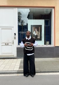 Una persona è in piedi davanti a un negozio, indossando un gilet a righe bianche e nere sopra una camicia bianca, con pantaloni neri larghi e scarpe nere.