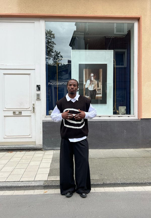 Een persoon staat voor een winkel, gekleed in een zwart-wit patroonvest over een witte blouse, met losse zwarte broeken en zwarte schoenen.