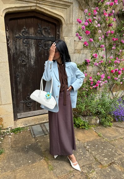 Blazer azul claro sobre un vestido marrón, bolso blanco con un detalle de flor y tacones blancos puntiagudos, con un muro de piedra floral de fondo.