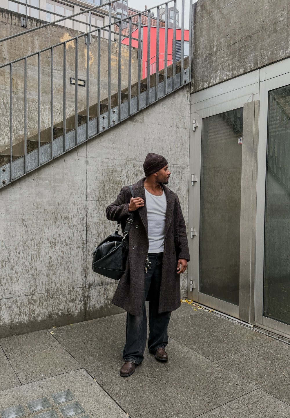 Dark wool coat over a white shirt, black trousers, and brown shoes. Carrying a black duffel bag, standing near glass doors and a concrete wall.
