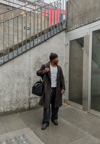 Dark wool coat over a white shirt, black trousers, and brown shoes. Carrying a black duffel bag, standing near glass doors and a concrete wall.
