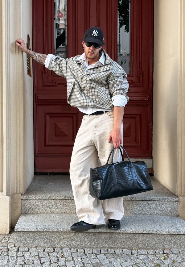 Hombre con chaqueta de cuadros, pantalones blancos, gorra negra, gafas de sol sujetando una bolsa negra, apoyado en una pared junto a los escalones de una puerta roja.