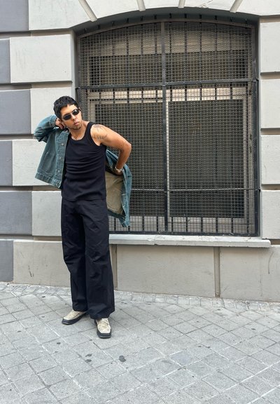 Top negro sin mangas, chaqueta de mezclilla, pantalones anchos negros y zapatos beige. El entorno presenta una pared texturizada y una ventana de rejilla metálica.