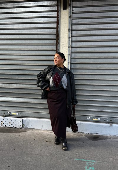 Persona con chaqueta de cuero oscura, suéter de rombos, falda larga y botas, de pie con bolso cerca de persianas metálicas cerradas en una acera de la ciudad.