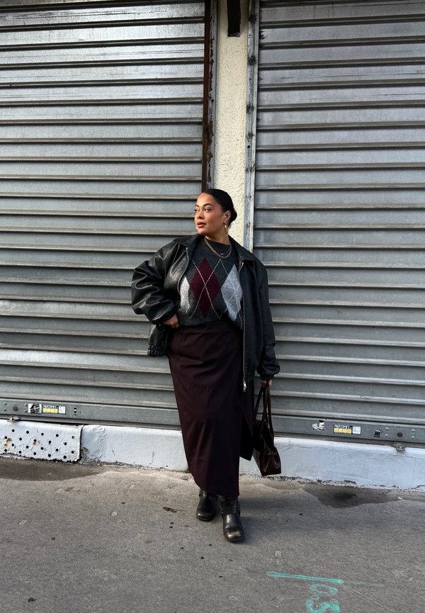 Osoba v tmavej koženej bunde, argyle svetri, dlhej sukni a čižmách stojaca s kabelkou pri zatvorených kovových roletách na mestskom chodníku.