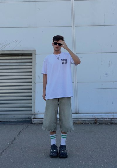 Una persona con una camiseta blanca de gran tamaño con texto negro, pantalones cortos de mezclilla con un toque verde, medias blancas a rayas y zuecos negros, de pie al aire libre.