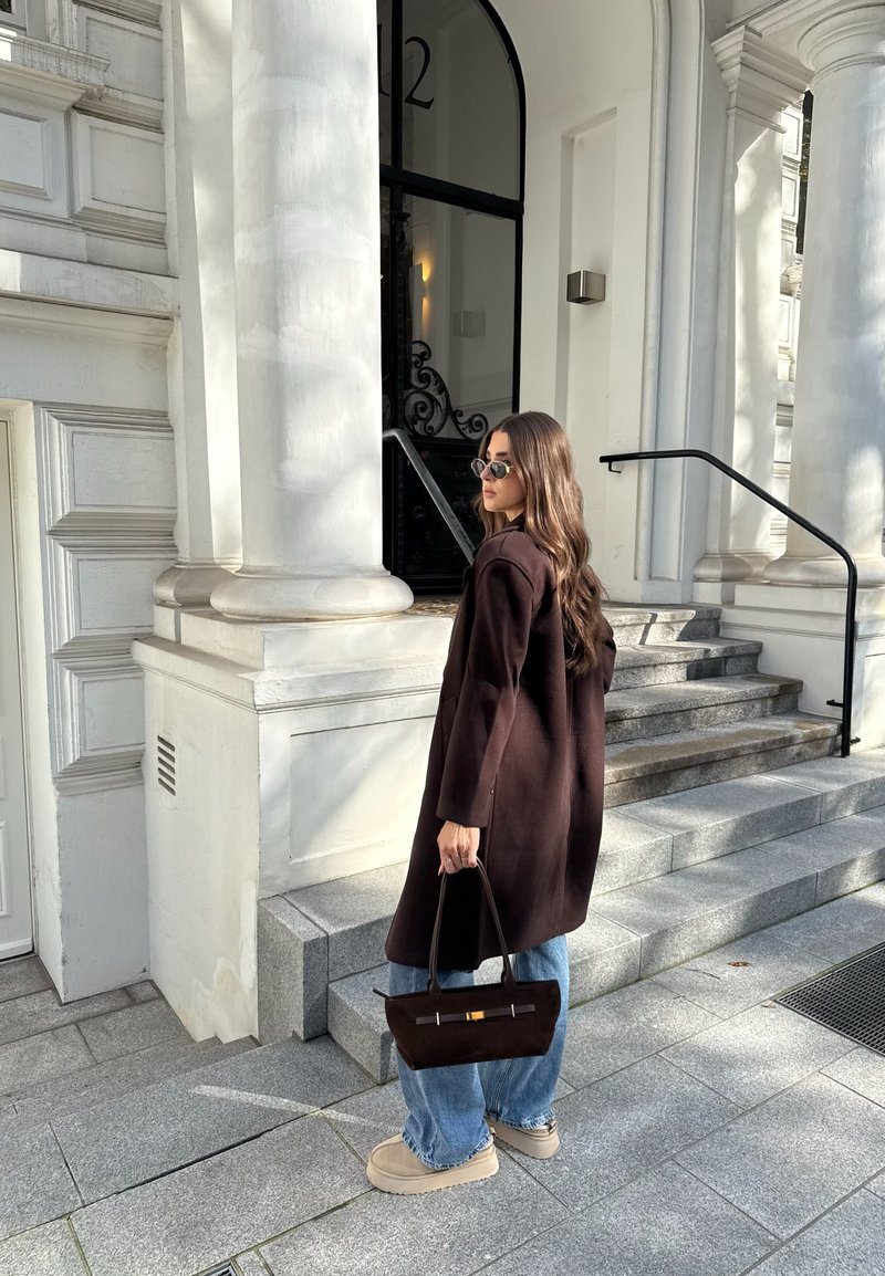 Brown wool coat, light blue loose-fitting jeans, beige platform shoes, holding a dark brown handbag with gold accents, standing on grey stone steps.