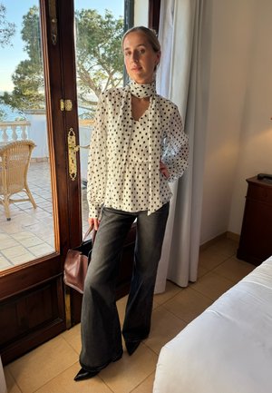 Balta palaidinė su baltais taškais, V formos kaklu ir klostėmis, derinama su tamsiai pilkais, aukštu liemeniu platėjančiais džinsais ir juodais smailiais bateliais. Ruda rankinė.