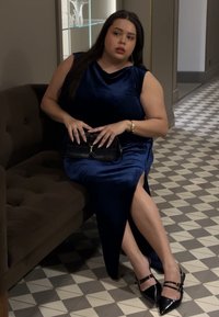 Aksamitna niebieska sukienka z bocznym rozcięciem, siedząca na brązowej pikowanej kanapie. Czarna torebka i czarne szpilki z paskami. Geometryczna podłoga z płytek.