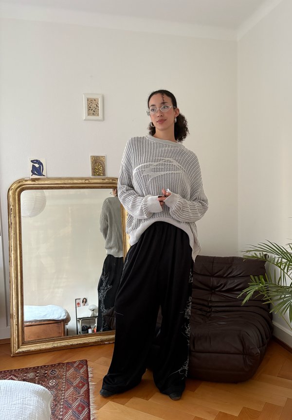 Personne portant des lunettes, un pull gris ample et un pantalon noir large, se tient à côté d'un grand miroir orné dans une pièce minimaliste.