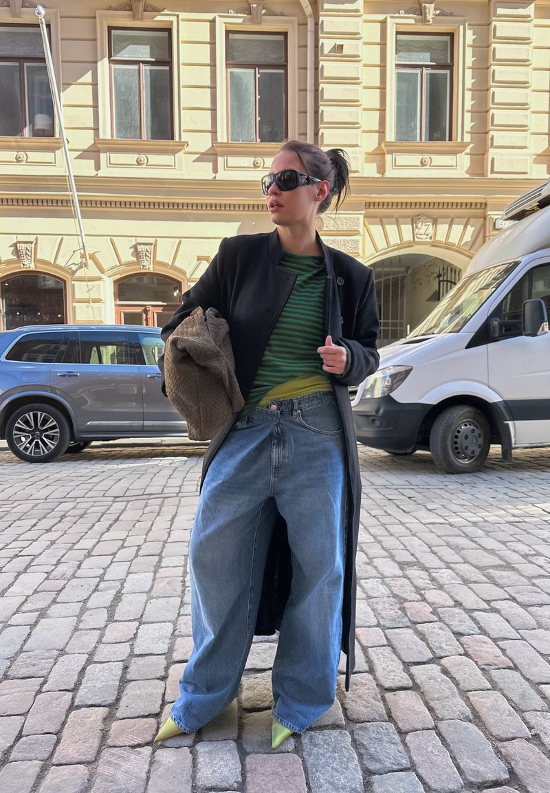 Femme portant un jean bleu surdimensionné, un long manteau noir, une chemise rayée verte, des chaussures pointues jaunes et des lunettes de soleil dans une rue pavée.