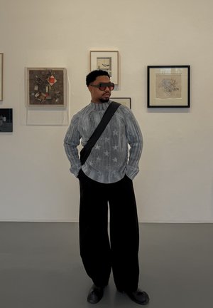 Hombre con gafas de sol, suéter gris con estampado de estrellas y pantalones negros de pierna ancha, de pie en una galería de arte con obras enmarcadas en la pared.