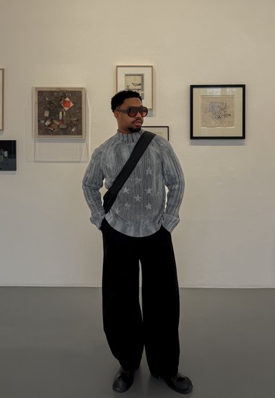 Hombre con gafas de sol, suéter gris con estampado de estrellas y pantalones negros de pierna ancha, de pie en una galería de arte con obras enmarcadas en la pared.