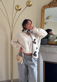Pull court en tricot blanc, pantalon gris à jambes larges et écharpe à motifs noir et blanc. Petit sac à main beige et lampe sur pied dorée en arrière-plan.
