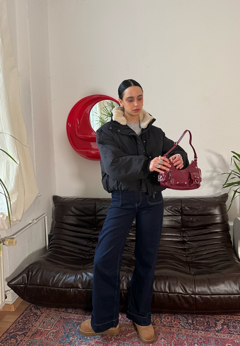 Doudoune noire avec un col en fausse fourrure, jeans bleu foncé à jambes larges, et sac à main bordeaux brillant. Chaussures marron et miroir décoratif rouge.