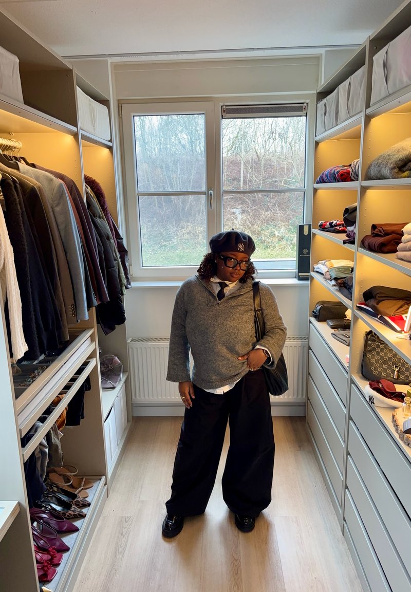 Person iført grå sweater, sorte vide bukser, baret og briller, stående i et godt oplyst walk-in-closet med tøj og sko på hylder.