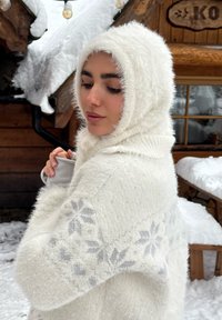 Fehér, bolyhos kötött kendő hozzáillő kapucnival, enyhe szürke hópehely mintával a ujjain, havas háttér előtt.