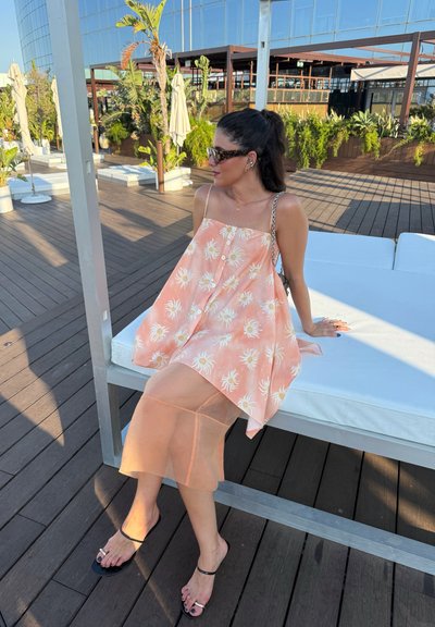 Vestido floral de durazno con detalles de botones, superpuesto a una falda sheer de durazno. Sandalias negras con tiras. Al aire libre sobre una cubierta de madera, en un entorno soleado.