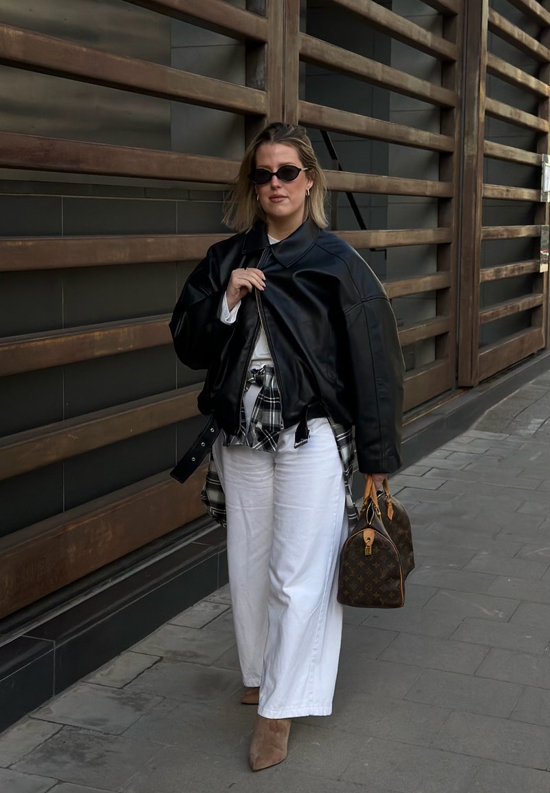 Femme portant une veste en cuir noire, un pantalon large blanc, une chemise à carreaux nouée autour de la taille, des lunettes de soleil, tenant un sac à main marron à motifs, marchant dans la rue.