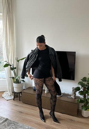 Chaqueta de cuero negro, top negro ajustado y medias negras con estampado de encaje. Tacones de aguja completan el conjunto. Ambiente interior con plantas.
