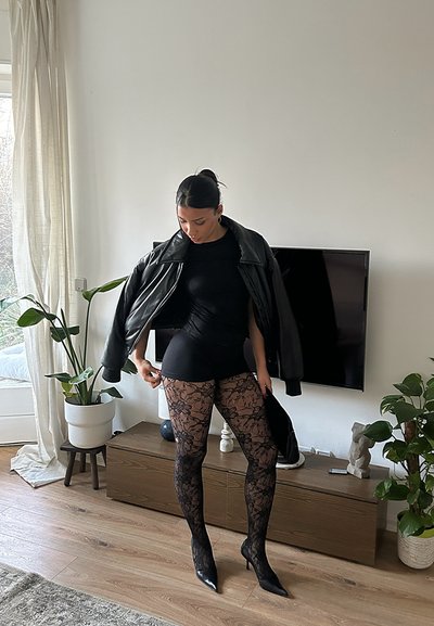 Chaqueta de cuero negro, top negro ajustado y medias negras con estampado de encaje. Tacones de aguja completan el conjunto. Ambiente interior con plantas.