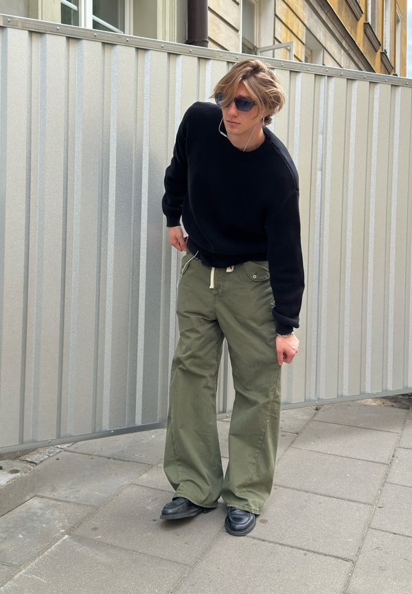 Jeune personne portant des lunettes de soleil, un pull noir et un pantalon vert à jambes larges, debout sur un trottoir avec une clôture métallique et des bâtiments derrière.