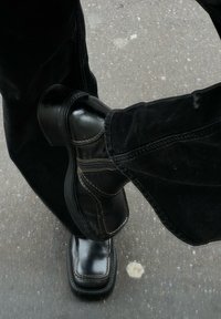 Μαύρες δερμάτινες μπότες αστραγάλου με τετράγωνη μύτη, έντονη ραφή και υφασμάτινες σόλες, φορεμένες με μαύρα παντελόνια σε υφασμάτινη επιφάνεια.