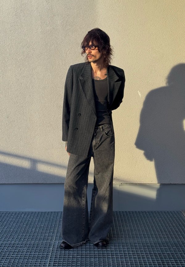 Homme aux cheveux mi-longs et lunettes, portant un blazer sombre à rayures, un débardeur noir et un jean gris à jambes larges, debout contre un mur beige.